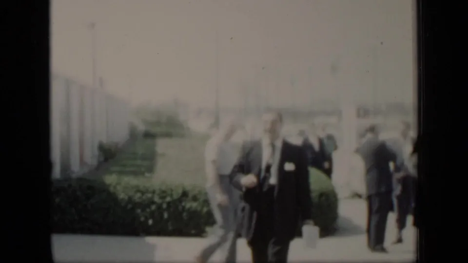 A suited man and other adults move through an outdoor New York 1948 setting beside hedges and low buildings.