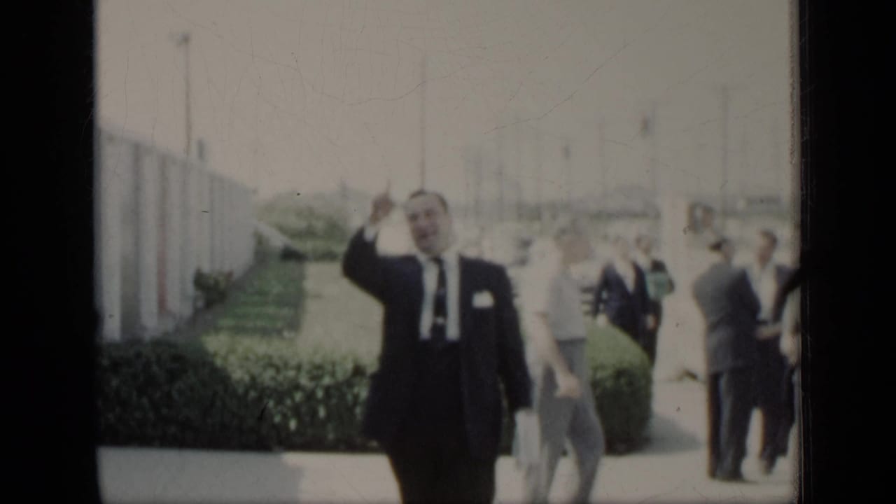Man in a dark suit waves while walking past hedges and pedestrians.