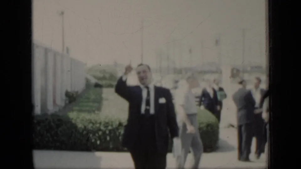 In 1948 New York home-movie footage, a man in a dark suit waves toward the camera beside hedges and passing adults.