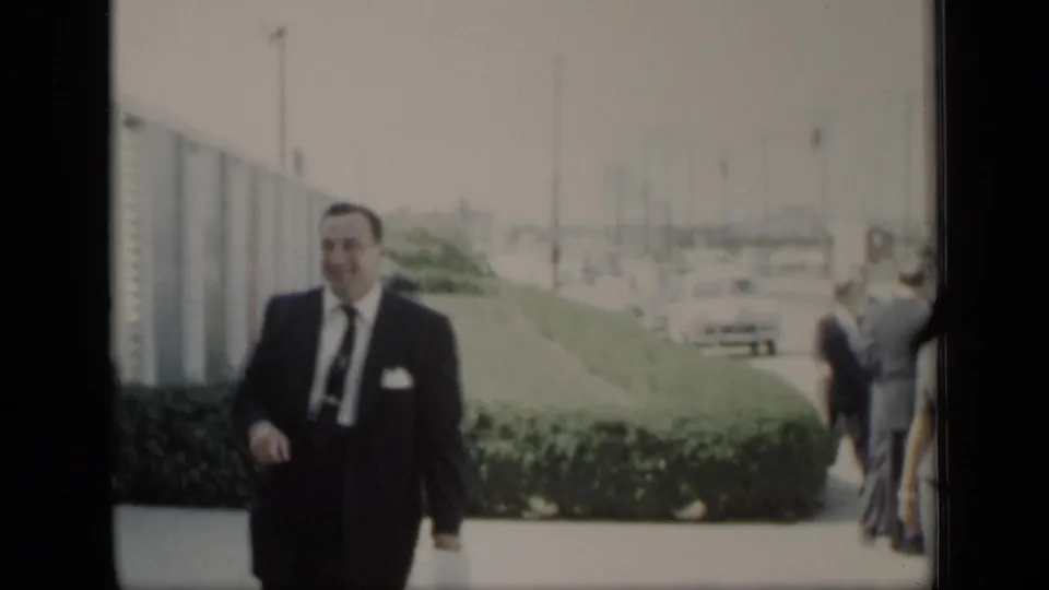 A suited man walks toward the camera in 1948 New York, with hedges, parked or moving cars, and other adults in the background.