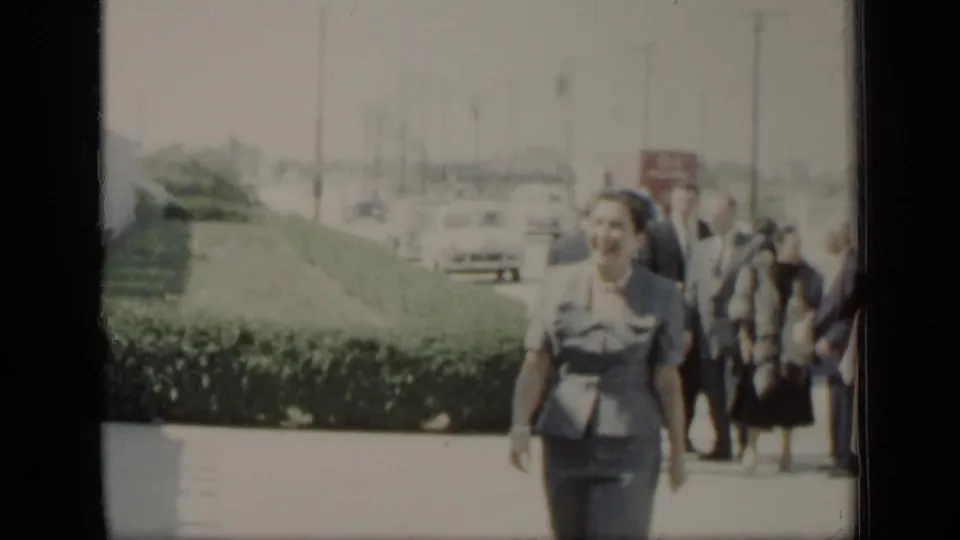 A woman in a blue suit walks toward the camera in the 1948 New York scene, with hedges, cars, and adults gathered behind her.