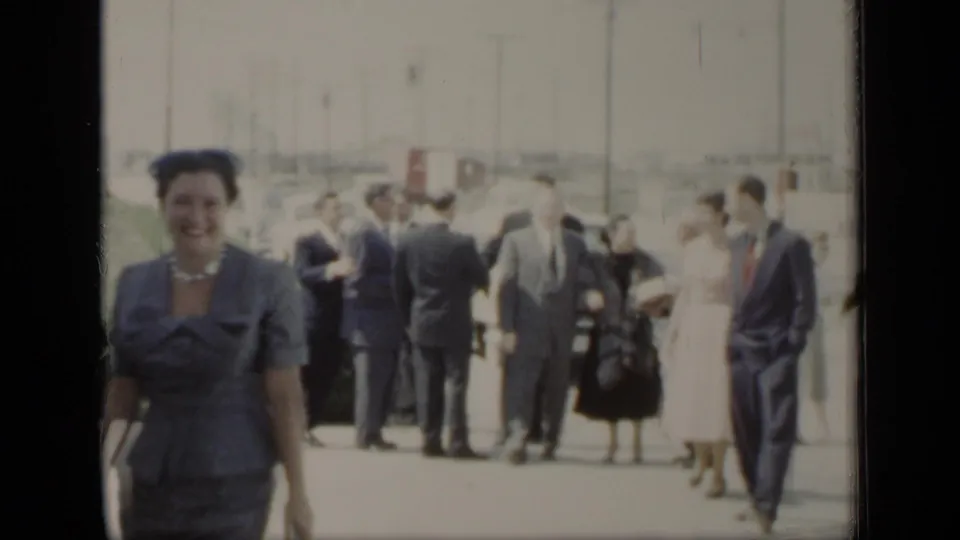 A smiling woman in a blue suit and necklace faces the camera in 1948 New York, with formally dressed pedestrians gathered behind her.