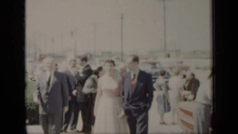 In 1948 New York home-movie footage, a bride and groom walk beside guests on a sunny path with cars and utility poles behind them.