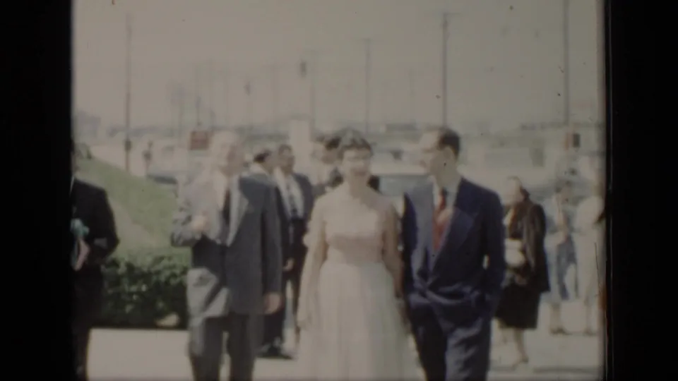 The bride and groom continue along the walkway in 1948 New York, with formally dressed guests, hedges, cars, and utility poles visible.