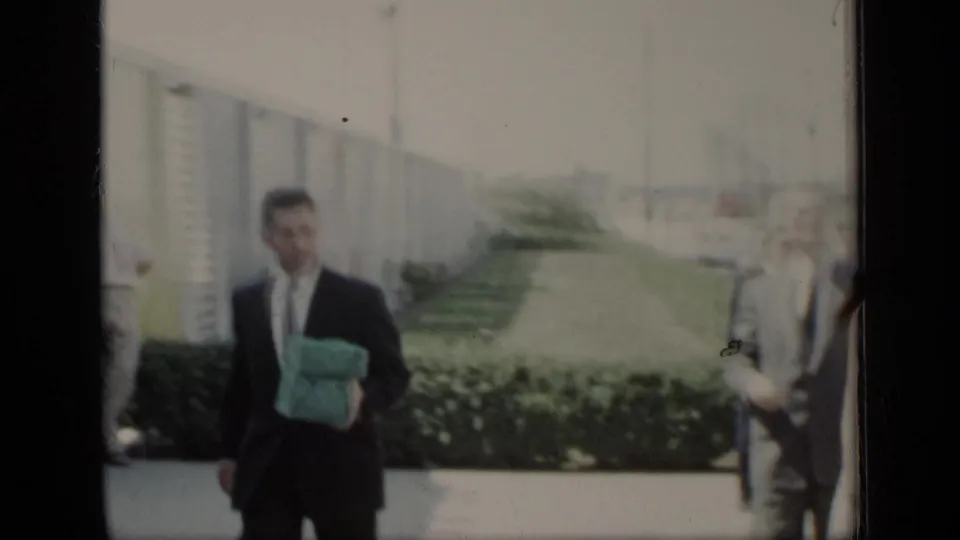 A suited man carrying a turquoise item walks near the hedge and building wall as other wedding guests move through the background.