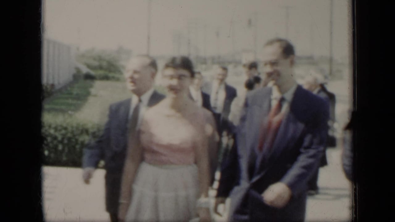 Three well-dressed adults walk along a bright sidewalk with hedges and people behind them.