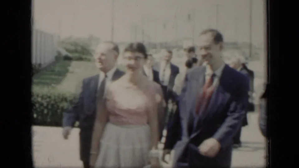 In 1948 New York home-movie footage, two men in suits and a woman in a pink top and pale skirt walk toward the camera on a sunlit sidewalk.