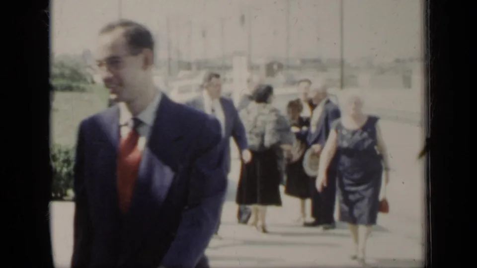 A suited man with a red tie passes close to the camera as other formally dressed adults walk behind him on a bright New York sidewalk.