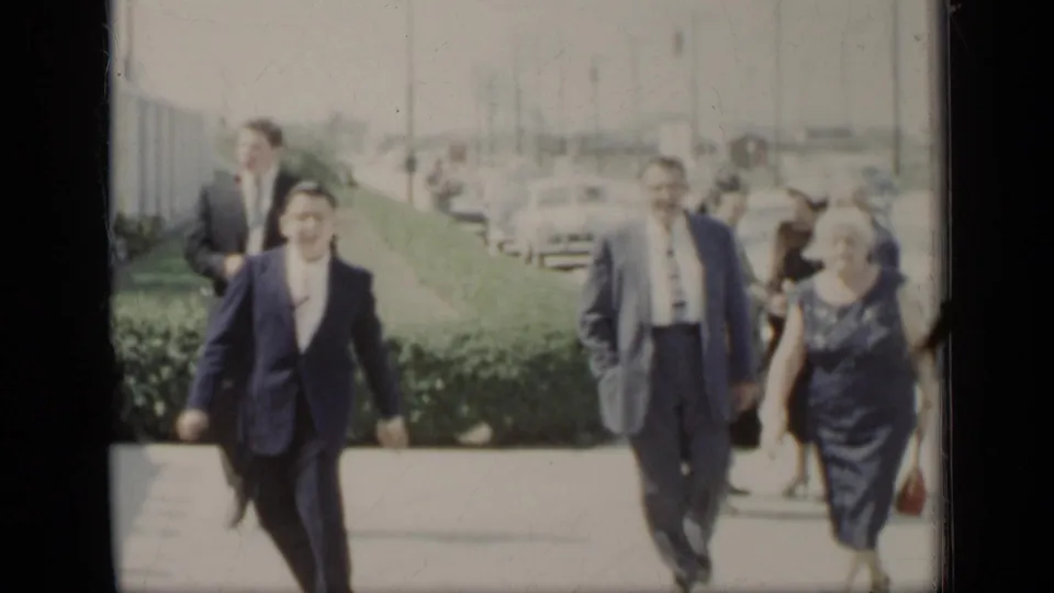 A group of adults in mid-century formal clothing walks toward the camera beside hedges, with several cars visible behind them in New York.