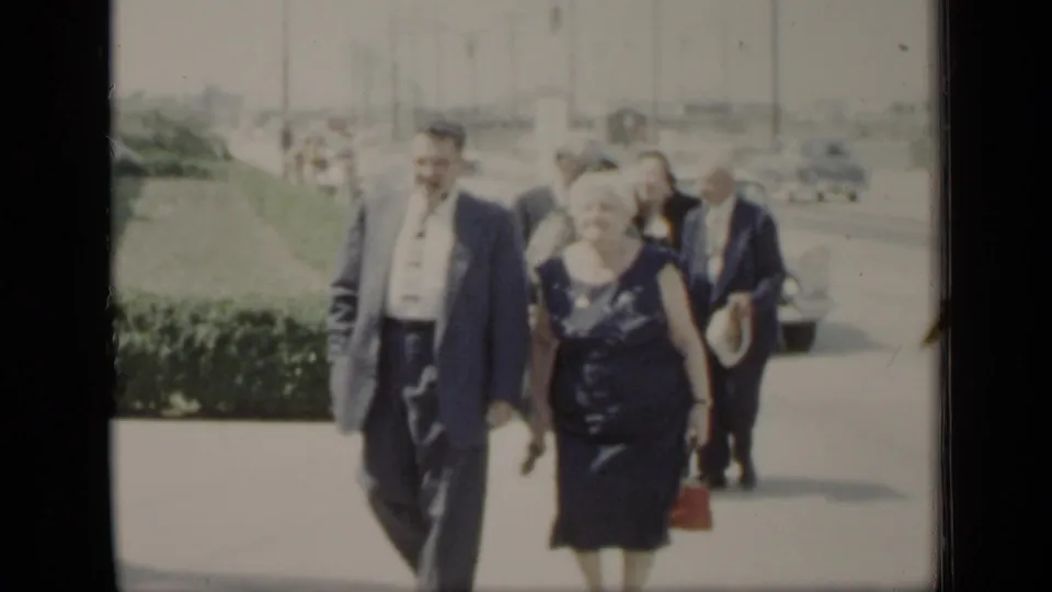 A man in a suit and a woman in a dark dress carrying a red purse walk forward as other adults follow on the sunlit New York sidewalk.