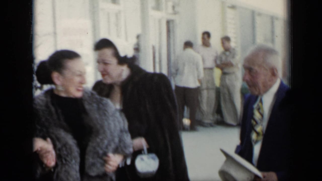 Older adults in formal clothing walking near a light-colored building.