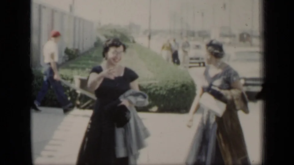 Two formally dressed women appear on a sunny walkway beside hedges, with a building wall and distant cars behind them.