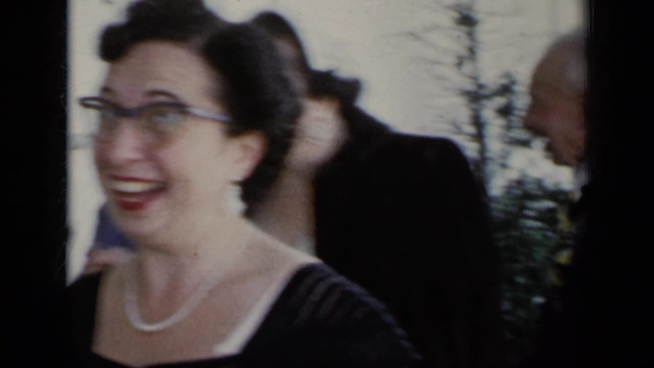 Woman in glasses smiles near other adults in a street-side home movie frame.