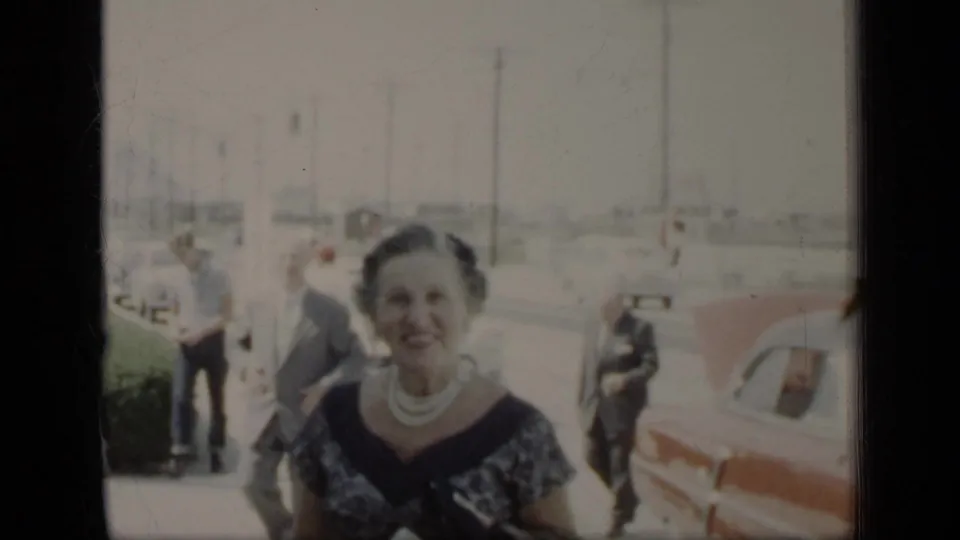 The woman in a dark patterned dress faces the camera near a sidewalk, parked car, and adults in 1948 New York footage.