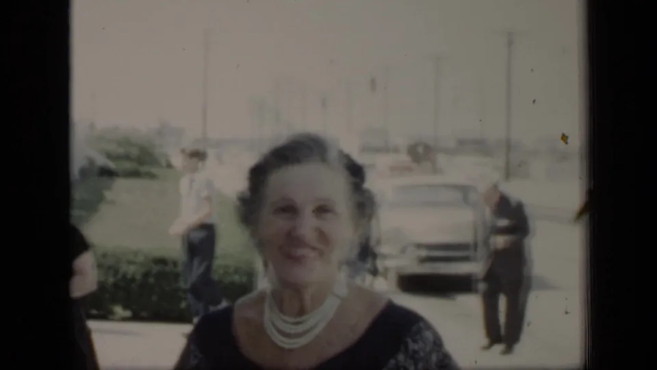 A smiling woman in a dark dress and pearls stands in front of a sidewalk, men, hedges, and a parked car in 1948 New York footage.