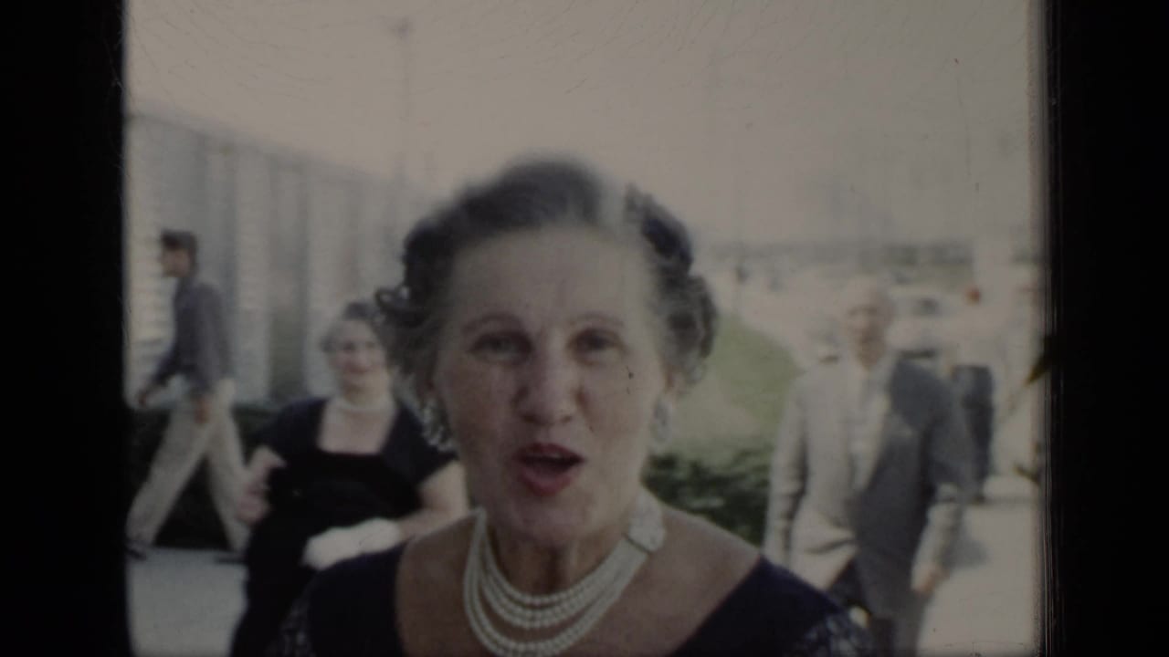 Woman in a dark dress and pearl necklace faces the camera as guests walk behind her.