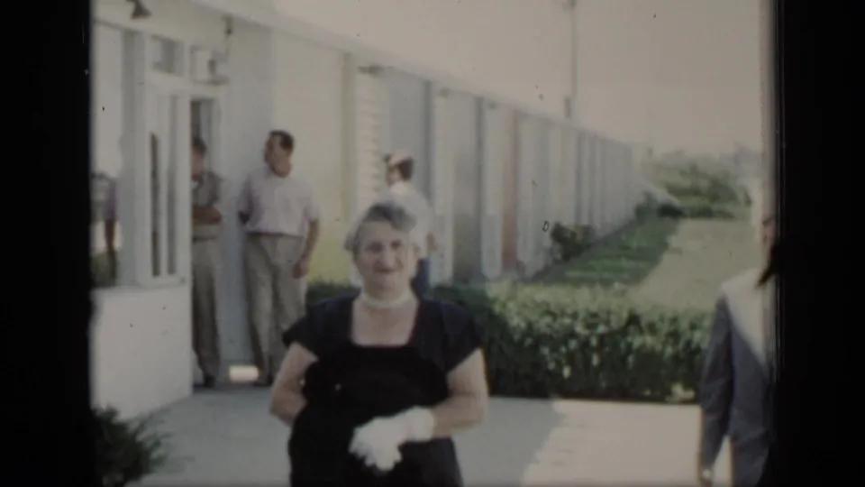 A woman in formal dress and white gloves stands outside a New York building as adults gather near the doorway in 1948 footage.
