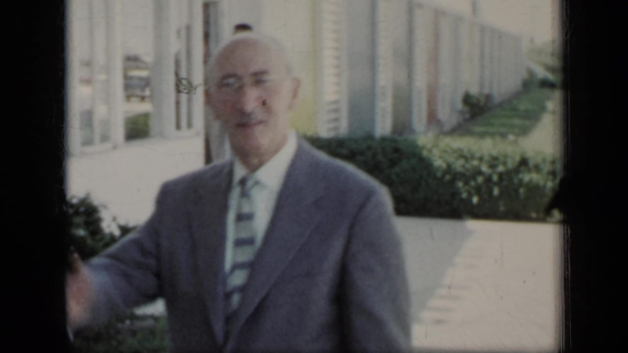 Older man in a suit smiles near the camera on a paved path.