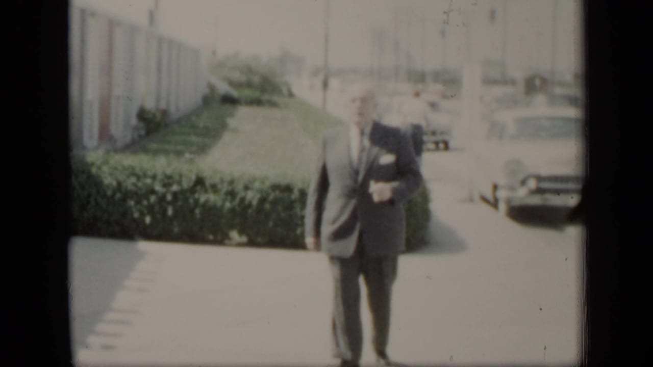 A man in a dark suit and tie walks toward the camera on a bright paved path, with hedges, a long building, and parked cars behind him.