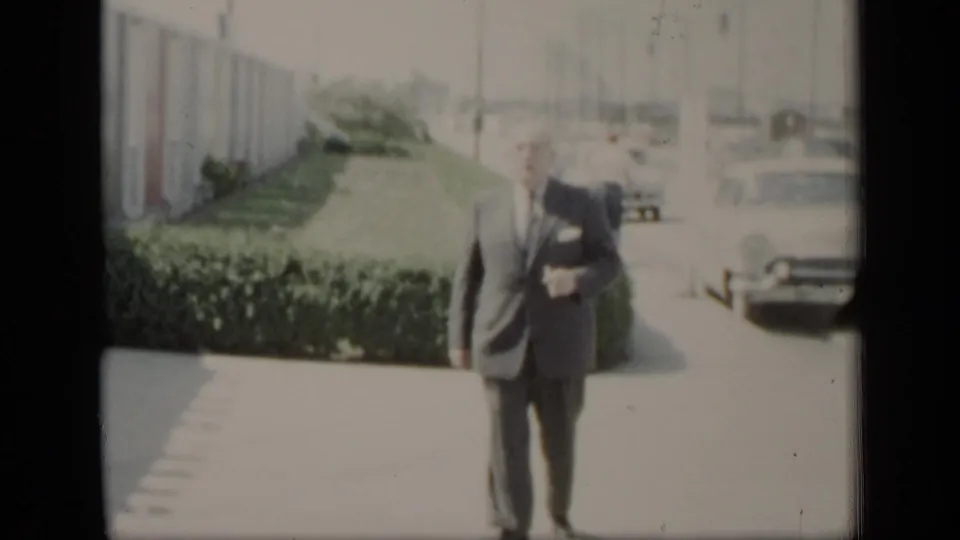 A man in a dark suit and tie walks toward the camera on a bright paved path, with hedges, a long building, and parked cars behind him.