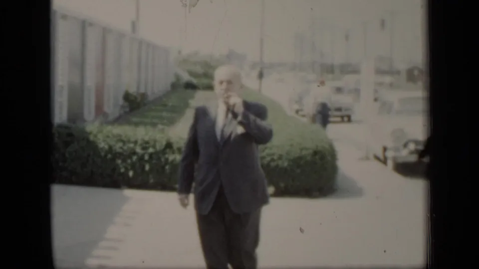 The suited man continues forward on the path and lifts a hand near his face, with the building, hedge, and parked cars still visible.
