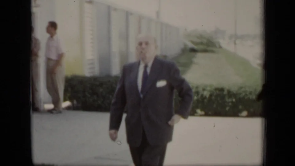 The suited man moves closer to the camera as another adult man stands by the building in the background near the hedges.