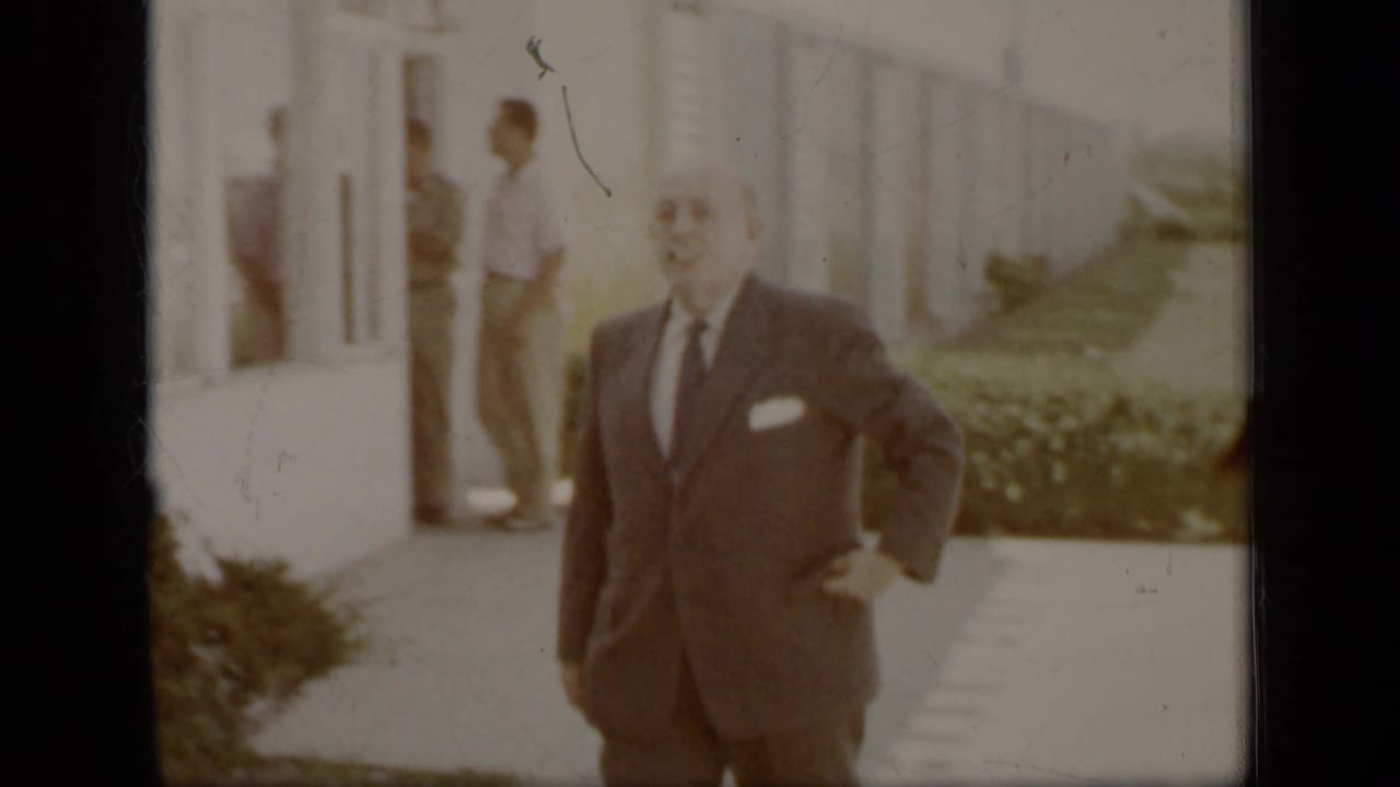 Suited man walking toward the camera outside a building, with another adult near a doorway.