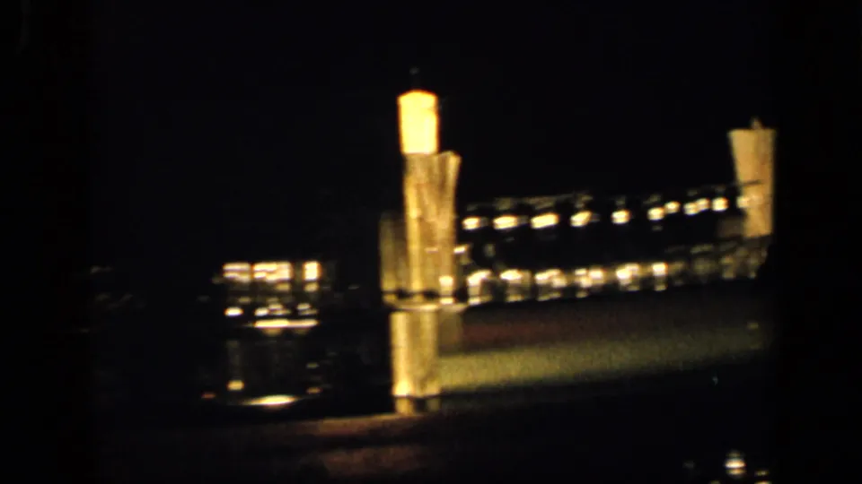 Blurry New York night exterior with bright illuminated structures and scattered lights, possibly near water or an industrial site.