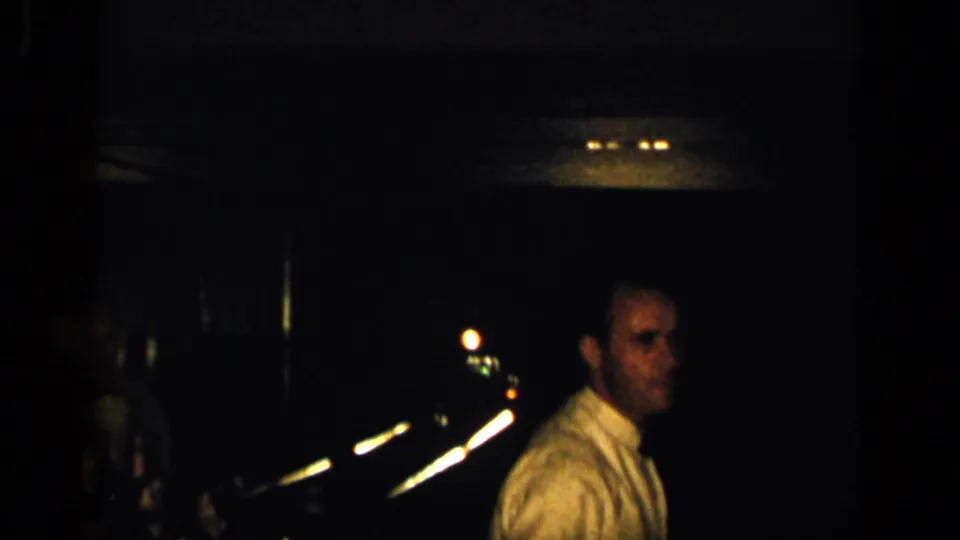 A man in a light shirt appears in a dim 1948 New York home-movie frame, with blurred night lights in the background.