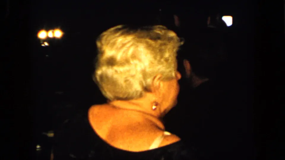 An older woman with short light hair is seen from behind during a dim 1948 New York night gathering with city lights nearby.