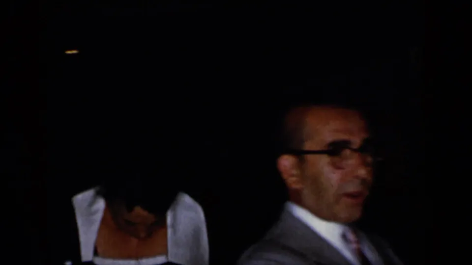 In dim 1948 New York home-movie footage, a suited man with glasses sits near a woman wearing a dark dress with a white collar.