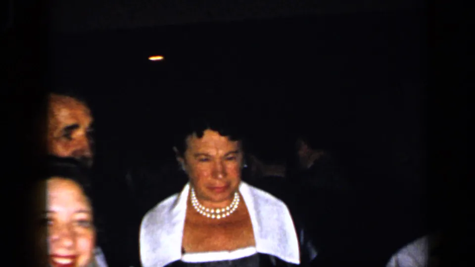 A small group of formally dressed adults appears in a dark interior, including a woman wearing pearls and a white-collared dress.