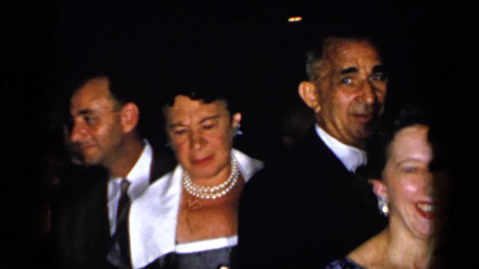 1948 New York party frame with a woman wearing pearl necklaces and a white-collared dress beside men in dark suits.