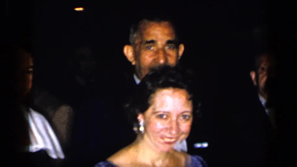 1948 New York party frame showing a smiling woman in a blue dress with men in dark suits behind her.