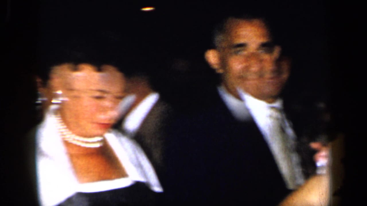 Formal guests in evening attire, including a woman with pearls and men in dark suits.