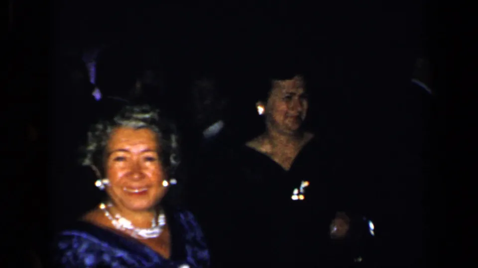 1948 New York home-movie frame with a smiling woman in formal dress and jewelry in a dark reception room.