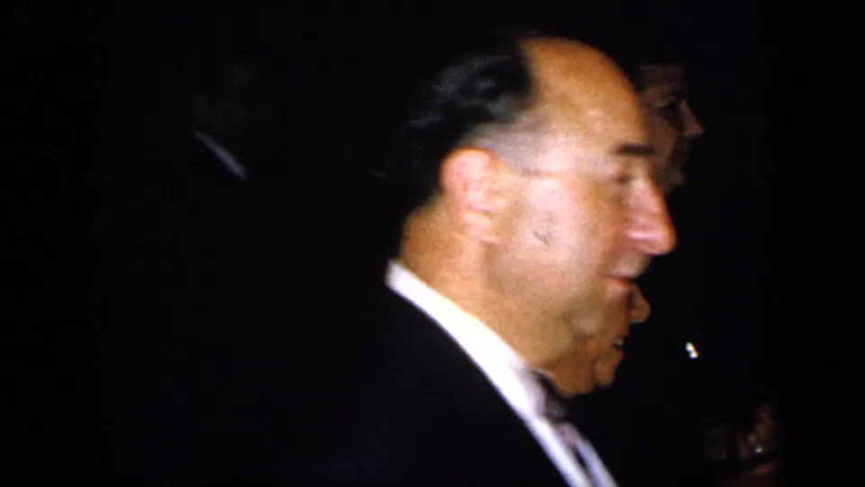1948 New York reception frame showing a suited man in profile amid shadowed formal guests.