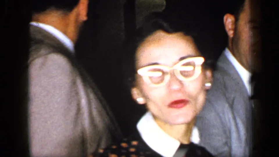 A woman in a dark formal dress turns toward the handheld camera while suited adults move through a dim New York corridor.