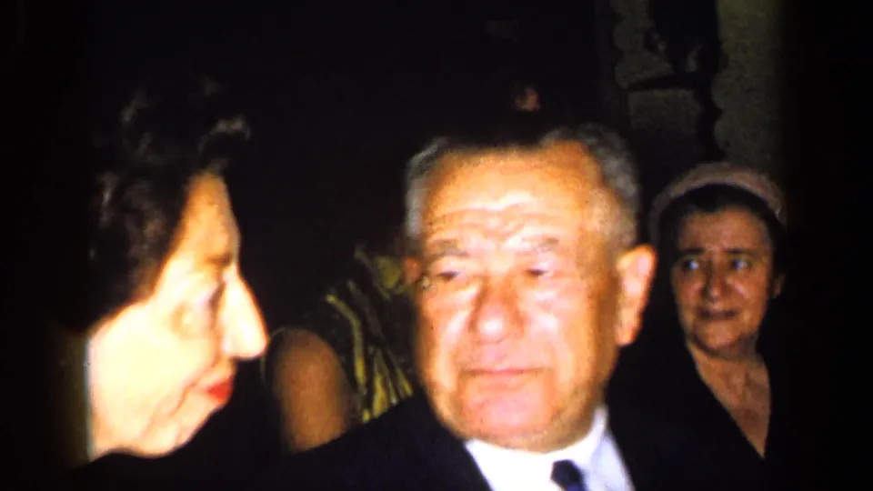 An older man in a dark suit and tie stands among women in formal clothing, lit by the handheld camera in 1948 New York home-movie footage.