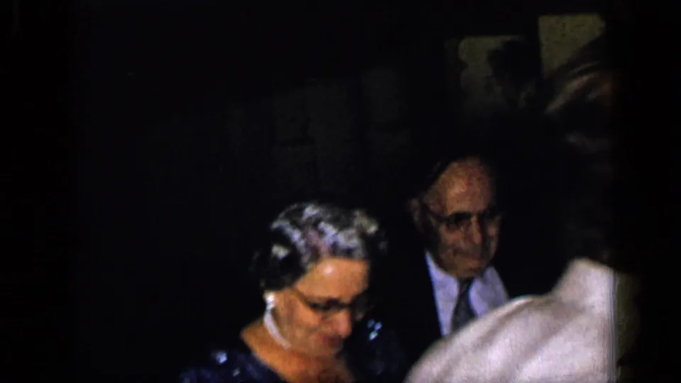 1948 New York frame with an older woman wearing glasses and a pearl necklace beside a suited man in low light.