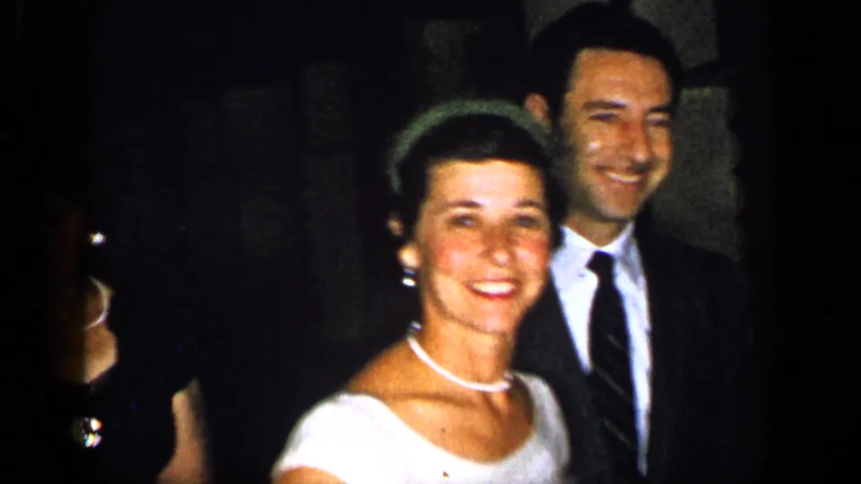 1948 New York home-movie frame showing a woman in a white formal dress and pearls with a smiling suited man behind her.