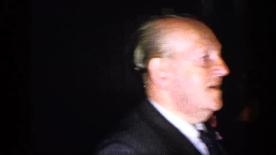 An adult man in a dark suit and tie moves past the camera during the 1948 New York formal indoor gathering.