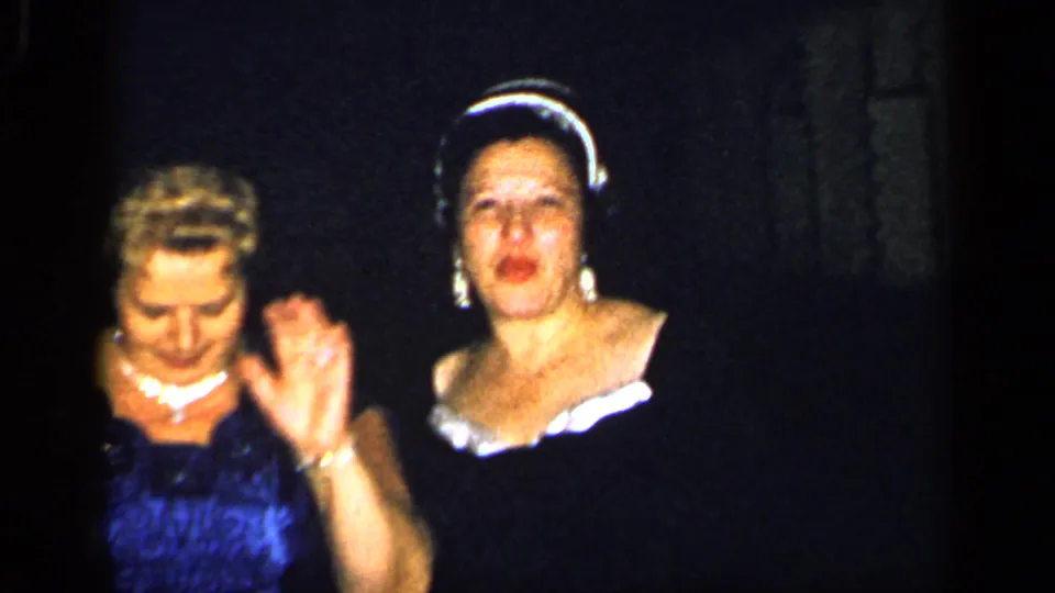 1948 New York home-movie frame with two women in formal dresses, including one in blue with pearls and one in a dark dress with a light collar.