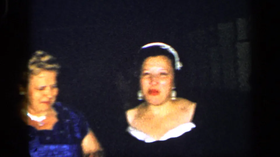 1948 New York 8mm frame showing two women in formal attire, one in a blue dress and pearls and another in a dark dress with a light neckline.