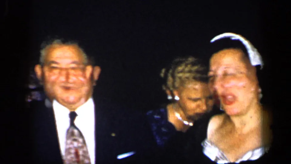 1948 New York home-movie frame with a smiling suited man and two women in formal dresses, one leaning forward near the camera.