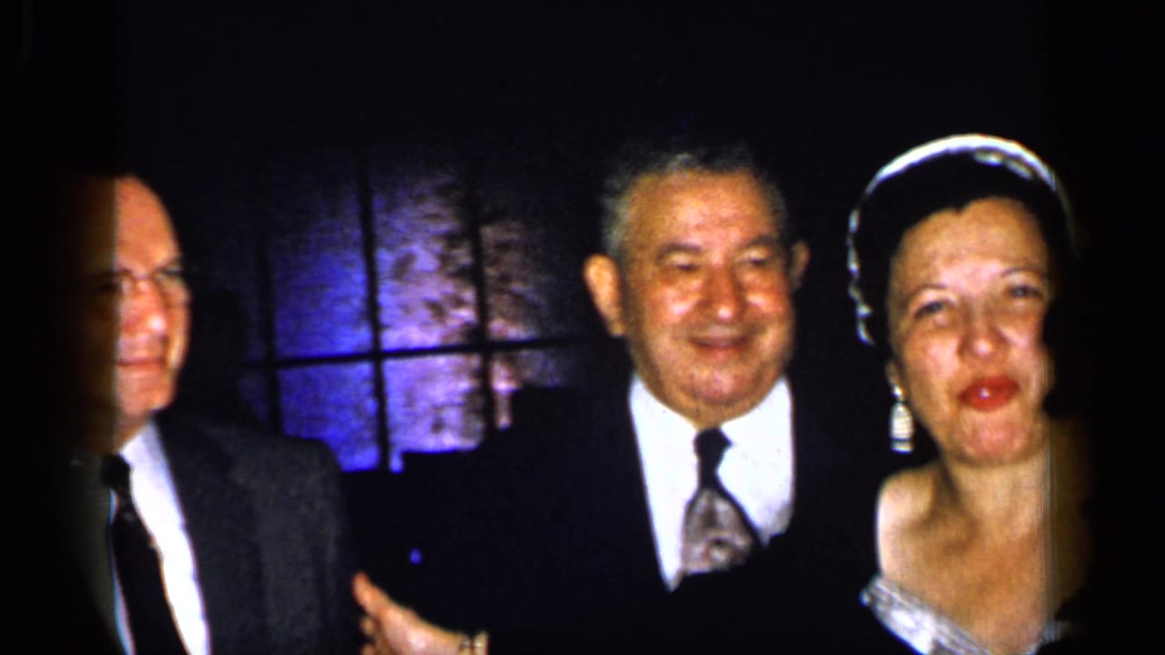 Formally dressed adults stand close together in dim light, including a smiling woman in a headpiece.