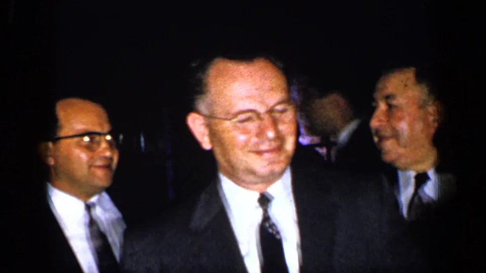 A 1948 New York 8mm frame shows men in suits beside a woman in formal dress and jewelry amid a crowded dim gathering.