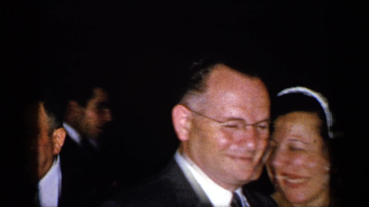 Smiling man in a suit beside a woman wearing a headpiece in a dark evening setting.