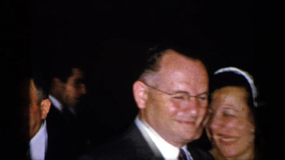 In 1948 New York home-movie footage, a man in a dark suit smiles beside a woman in formal dress with a light headpiece as other adults stand nearby in low light.