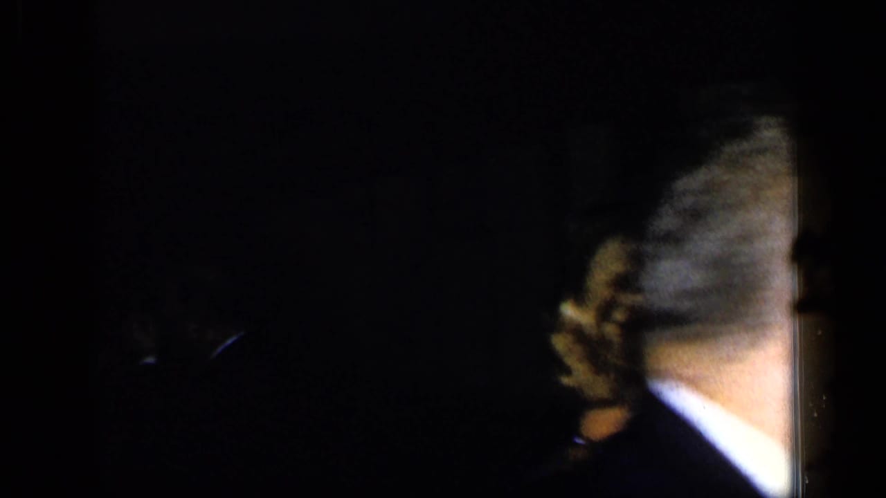 Dimly lit adults in formal wear with a man's shoulder and collar close to the camera.